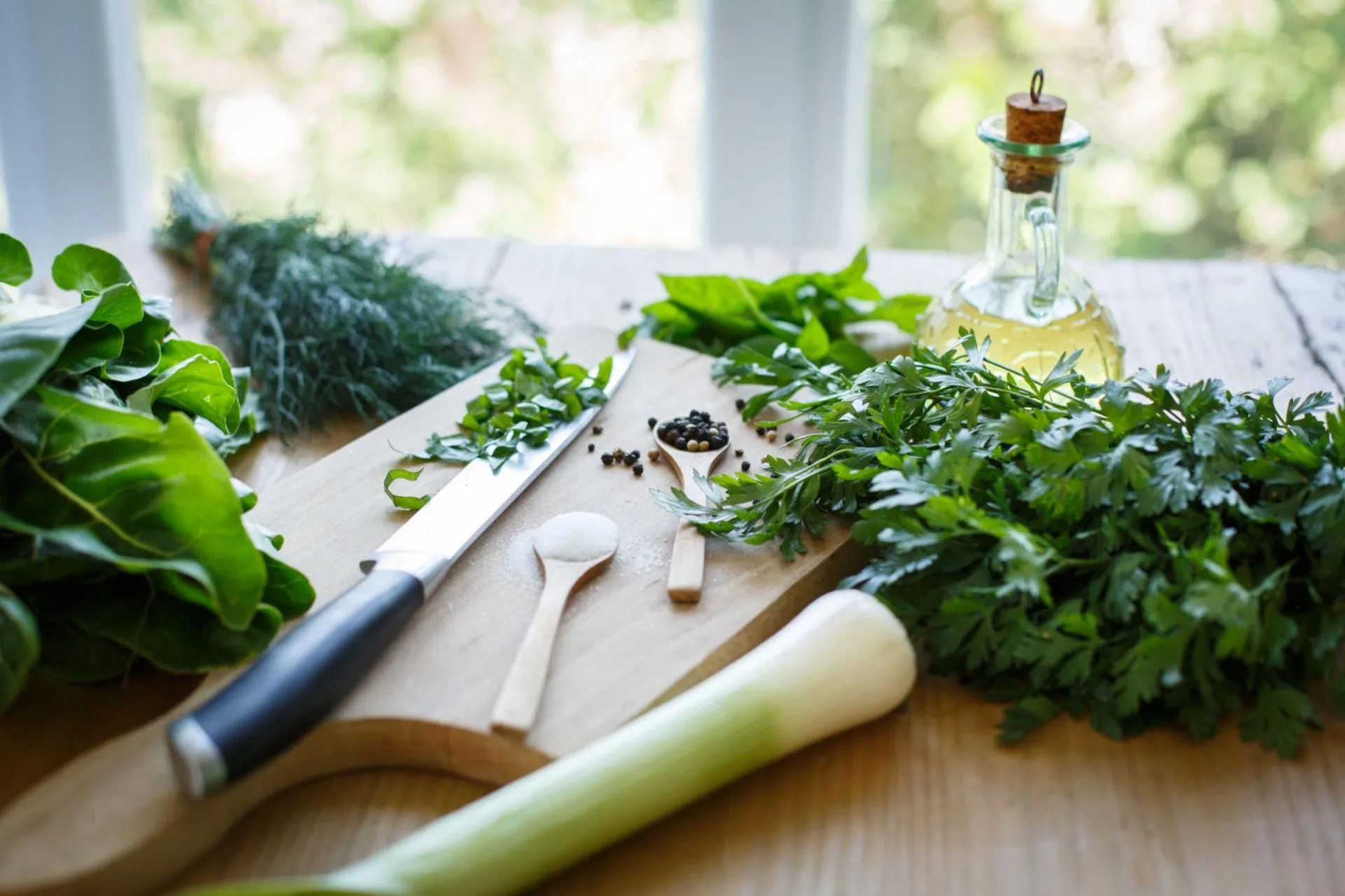 Fresh herbs, leek and cold-pressed olive oil arranged on a wooden board by a sunlit window