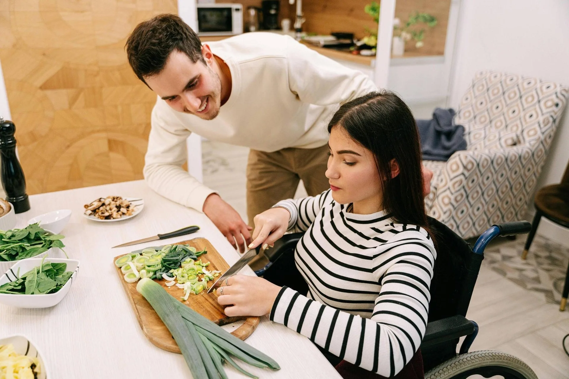 A young woman using a wheelchair preparing vegetables alongside her partner in a bright home kitchen