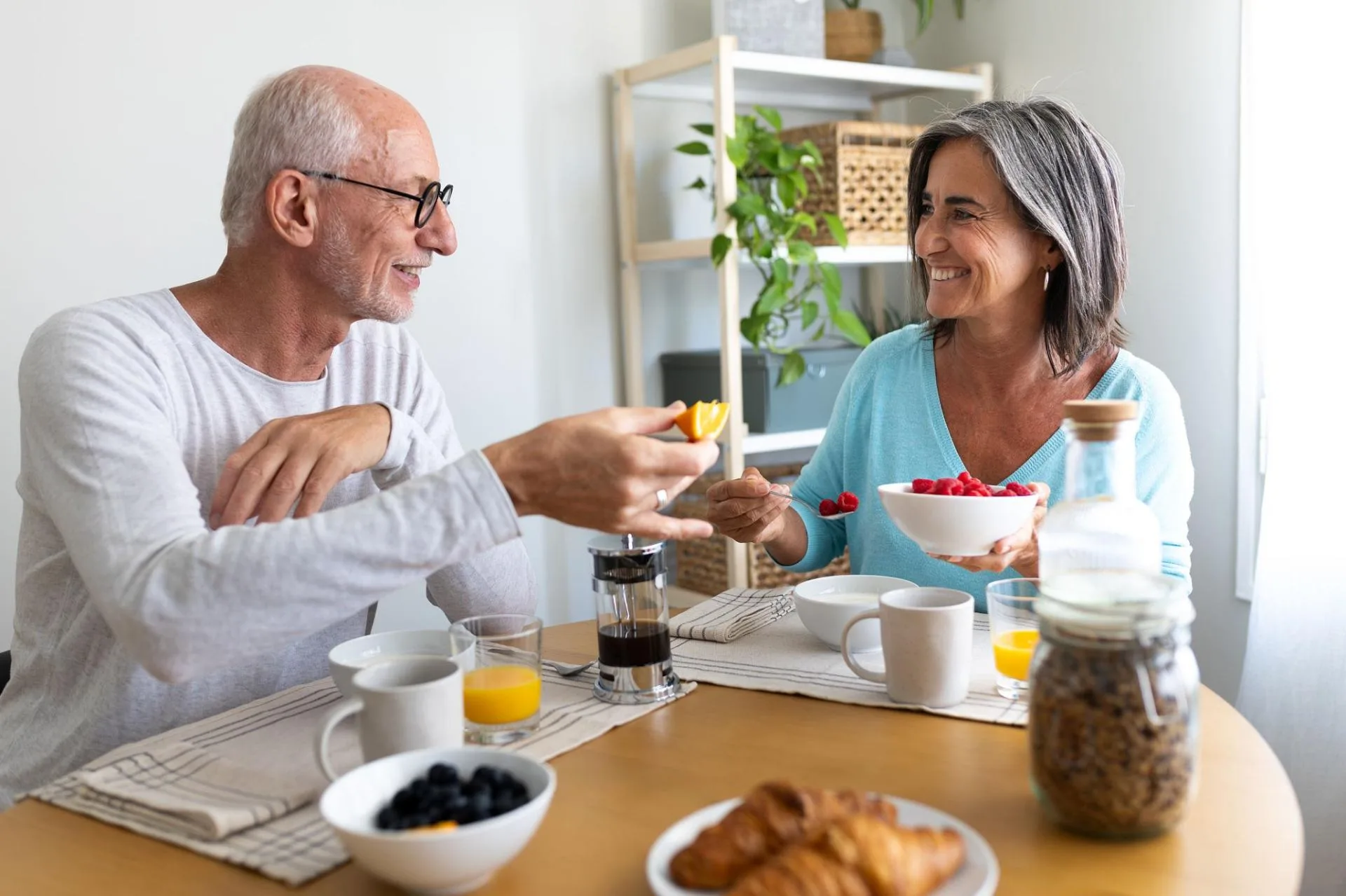 An older couple sharing breakfast at home together, smiling as they pass fresh fruit across the table