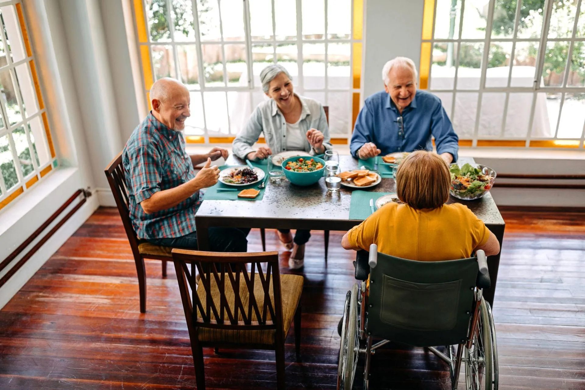 Four aged care residents enjoying a meal together at a dining table, one using a wheelchair, laughing in a bright communal dining room